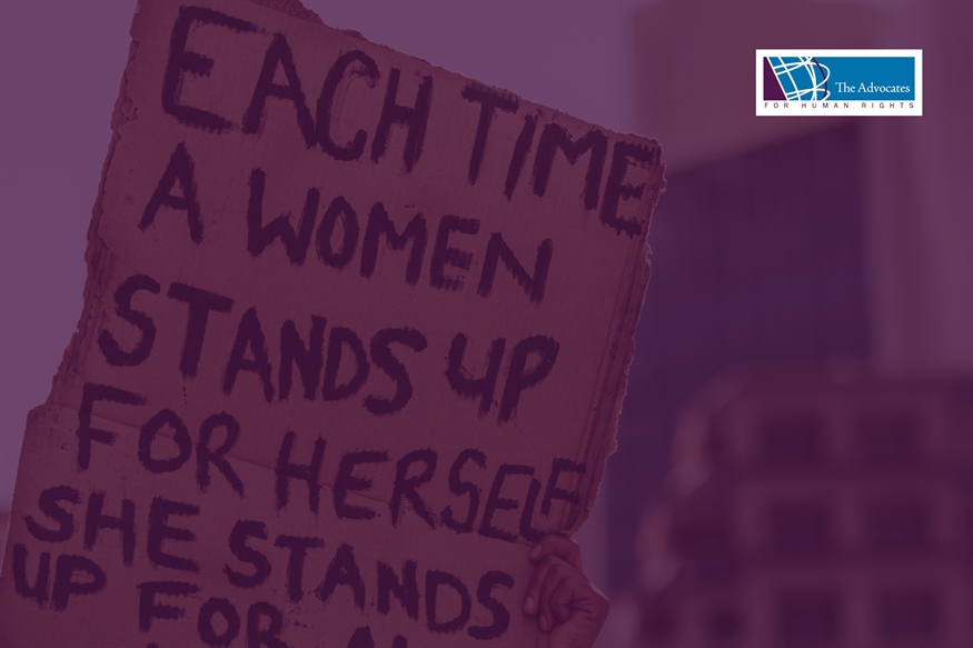 [ai] A person holding a handmade sign that reads, 'Each time a woman stands up for herself, she stands up for all.' The background features blurred buildings and a logo in the corner.